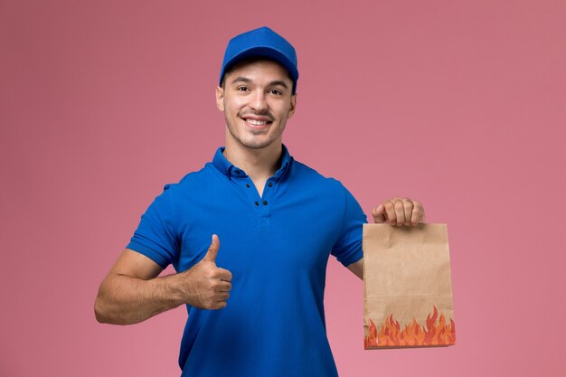 Vooraanzicht mannelijke koerier in blauw uniform met papieren voedselpakket met glimlach op de roze muur, uniforme dienstverlening