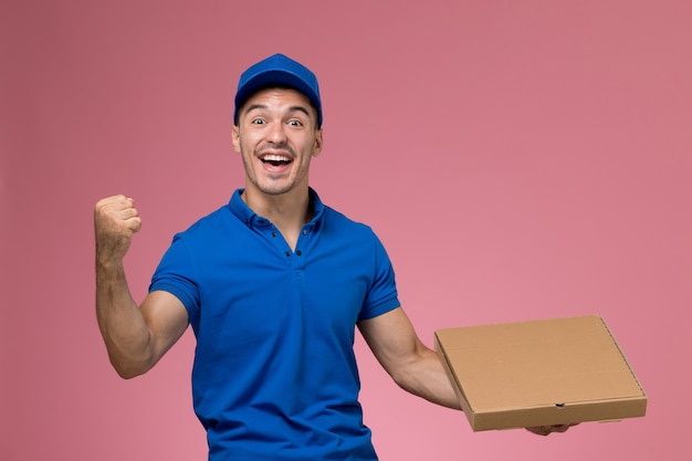 Vooraanzicht mannelijke koerier in blauw uniform met voedseldoos die zich verheugt op de roze muur, uniforme dienstverlening van de baanarbeider