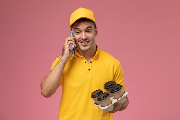 Gratis foto vooraanzicht mannelijke koerier in geel uniform bedrijf levering koffiekopjes praten aan de telefoon op roze bureau