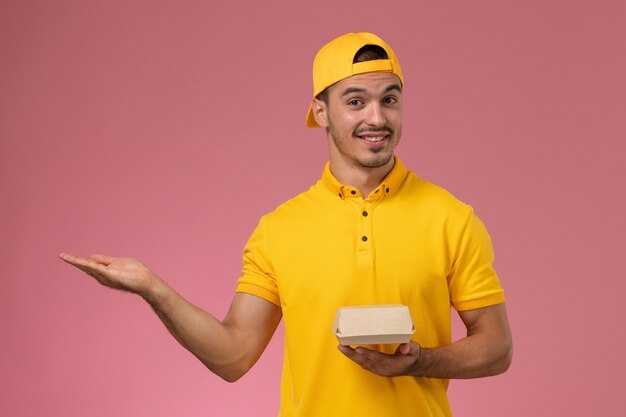 Vooraanzicht mannelijke koerier in geel uniform en cape die weinig pakket van het leveringsvoedsel op roze achtergrond houdt.