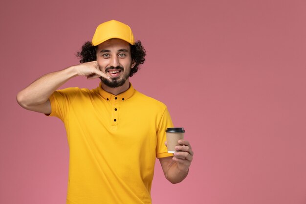 Vooraanzicht mannelijke koerier in geel uniform en cape met bezorging koffiekopje op lichtroze muur