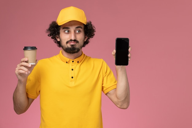 Vooraanzicht mannelijke koerier in geel uniform en cape met levering koffiekopje en telefoon op roze muur