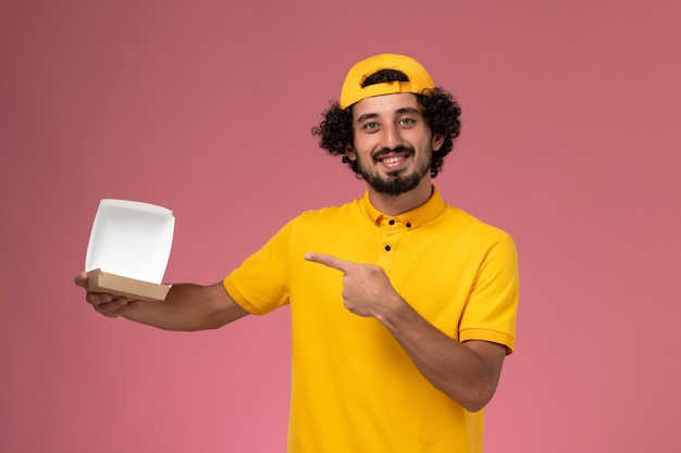 Vooraanzicht mannelijke koerier in geel uniform en cape met open klein pakket van het leveringsvoedsel op zijn handen op roze achtergrond.