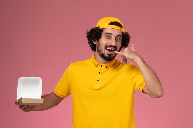 Vooraanzicht mannelijke koerier in geel uniform en cape met open klein pakket van het leveringsvoedsel op zijn handen op roze achtergrond.