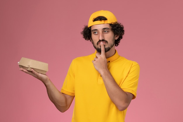 Vooraanzicht mannelijke koerier in geel uniform en cape met weinig bezorgvoedselpakket op zijn handen op de roze achtergrond.