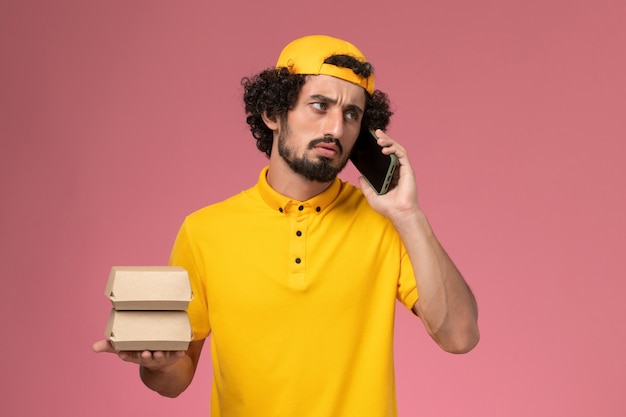 Vooraanzicht mannelijke koerier in gele uniforme cape met voedselpakketten op zijn handen praten aan de telefoon op lichtroze achtergrond.