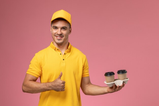 Vooraanzicht mannelijke koerier in gele uniforme koffiekopjes van de holdingslevering op de lichtroze achtergrond