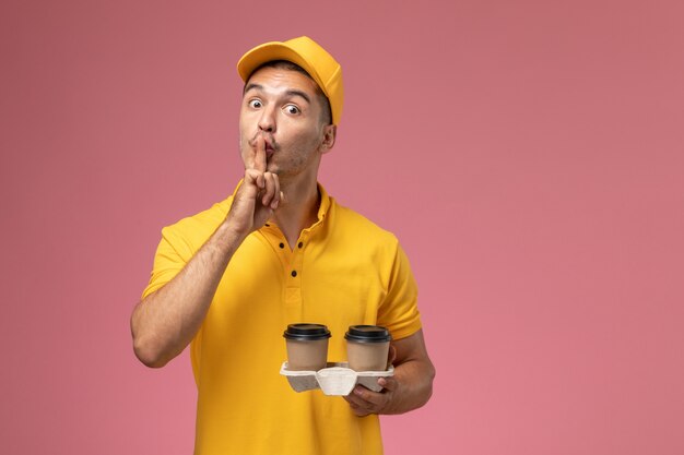 Vooraanzicht mannelijke koerier in gele uniforme koffiekopjes van de holdingslevering vragen stil te zijn op lichtroze achtergrond