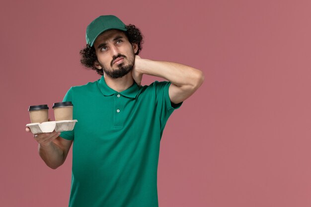 Vooraanzicht mannelijke koerier in groen uniform en cape bedrijf levering koffiekopjes denken op roze achtergrond service uniforme levering baan werk