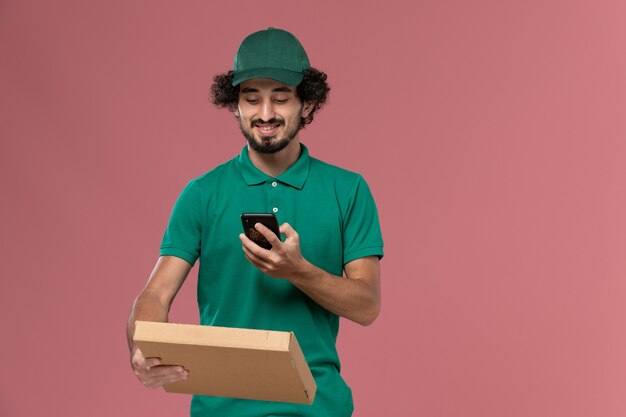 Vooraanzicht mannelijke koerier in groen uniform en cape bedrijf levering voedseldoos foto nemen op roze achtergrond service werknemer uniforme levering baan