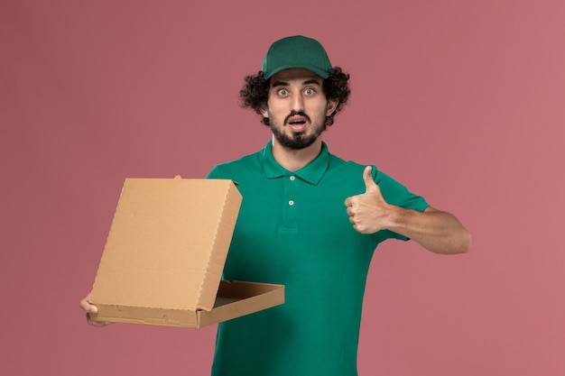 Vooraanzicht mannelijke koerier in groen uniform en cape bedrijf levering voedseldoos poseren op roze achtergrond service werknemer uniforme levering baan