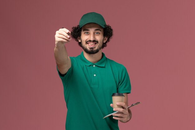 Vooraanzicht mannelijke koerier in groen uniform en cape met bezorging koffiekopje met blocnote en pen op het roze bureau uniforme bezorgdienst bedrijf baan