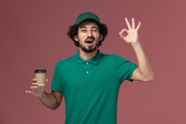 Vooraanzicht mannelijke koerier in groen uniform en cape met bezorging koffiekopje op de roze achtergrond uniforme bezorgdienst werk mannelijk bedrijf