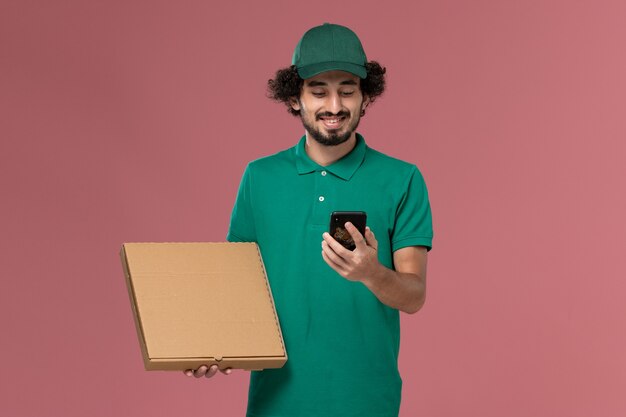 Vooraanzicht mannelijke koerier in groen uniform en cape met levering voedseldoos en zijn telefoon op lichtroze achtergrond service werknemer uniforme levering baan