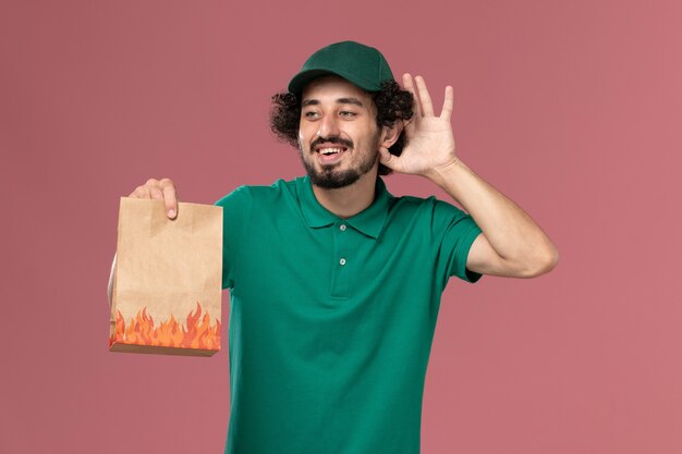 Vooraanzicht mannelijke koerier in groen uniform en cape met papier voedselpakket proberen te horen op de roze achtergrond service werknemer uniforme levering baan