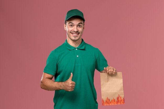 Vooraanzicht mannelijke koerier in groen uniform voedselpakket houden en glimlachend op de lichtroze achtergrond