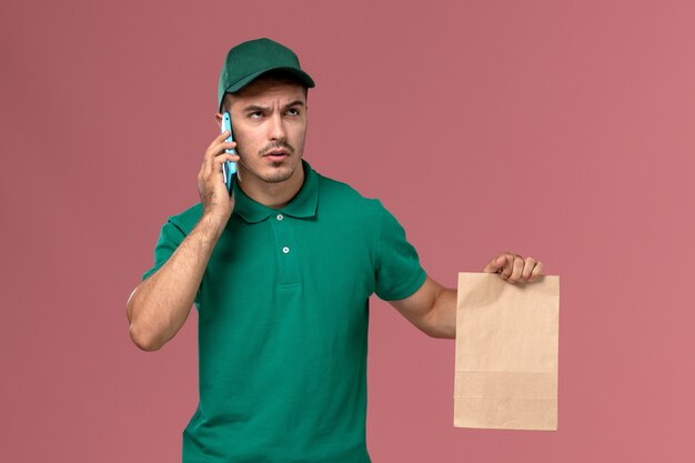 Vooraanzicht mannelijke koerier in groen uniform voedselpakket houden tijdens het praten aan de telefoon op roze achtergrond
