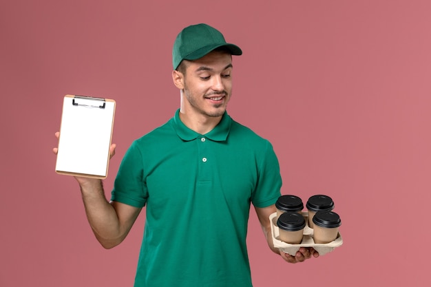 Vooraanzicht mannelijke koerier in groene uniform met bruine koffiekoppen blocnote op de roze achtergrond