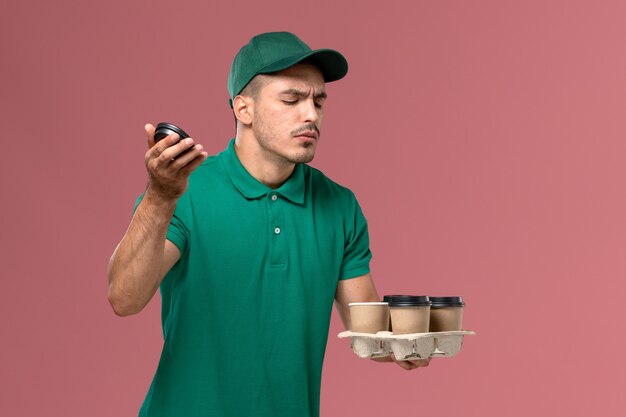 Vooraanzicht mannelijke koerier in groene uniforme de koffiekopjes die van de holdingslevering op de roze achtergrond ruiken