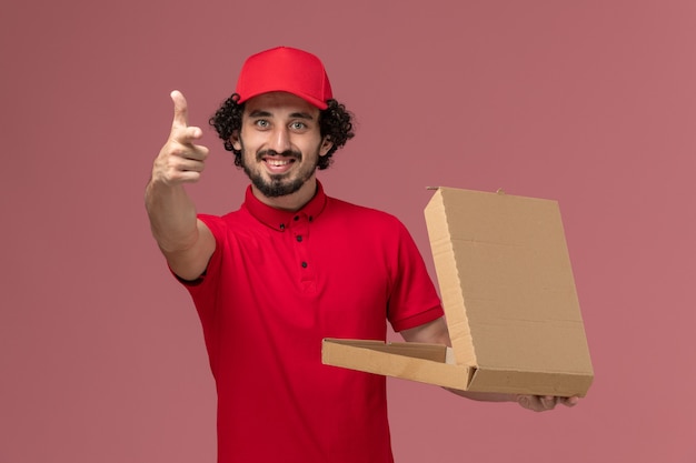 Vooraanzicht mannelijke koerier in rood overhemd en cape die lege doos van het leveringsvoedsel houden en op de roze werknemer van het de dienstleveringsbedrijf glimlachen