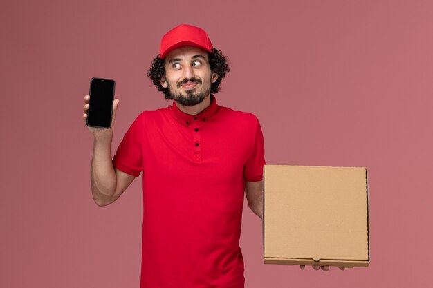 Vooraanzicht mannelijke koerier in rood overhemd en cape die lege doos van het leveringsvoedsel met telefoon op roze muur houden