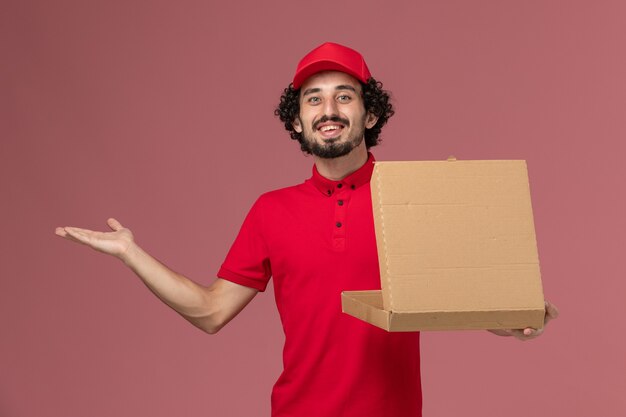 Vooraanzicht mannelijke koerier in rood overhemd en cape die voedseldoos op roze muur houden