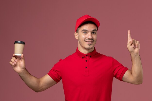 Vooraanzicht mannelijke koerier in rood uniform de koffiekop die van de holdingslevering op lichtroze muur glimlacht