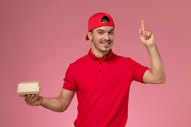 Vooraanzicht mannelijke koerier in rood uniform en cape die weinig leveringspakket met glimlach op roze achtergrond houdt.