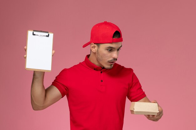 Vooraanzicht mannelijke koerier in rood uniform en cape met klein afleverpakket met blocnote op de roze achtergrond.