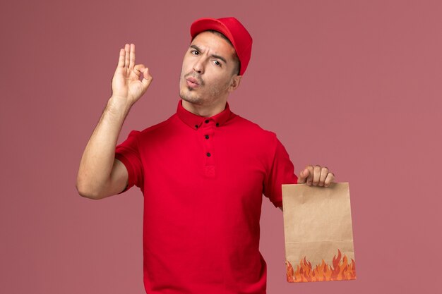 Vooraanzicht mannelijke koerier in rood uniform en cape met papier voedselpakket op roze vloerarbeider