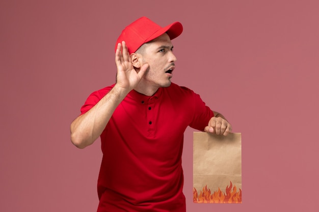 Vooraanzicht mannelijke koerier in rood uniform en cape met papieren voedselpakket en probeer te horen op de roze muurwerker