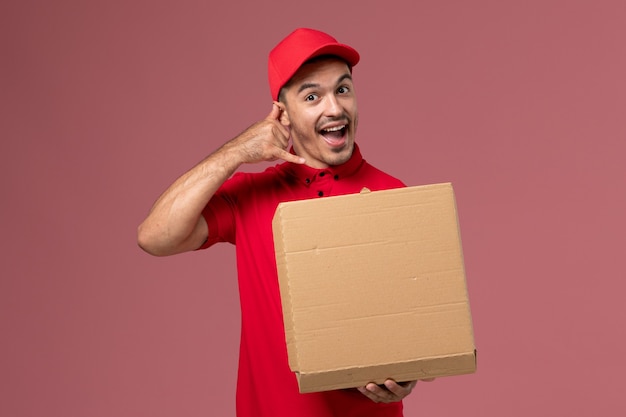 Vooraanzicht mannelijke koerier in rood uniform en cape met voedseldoos op de lichtroze muurarbeider