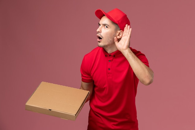 Vooraanzicht mannelijke koerier in rood uniform en cape met voedselleveringsdoos die probeert te horen op roze muurarbeiderbaan