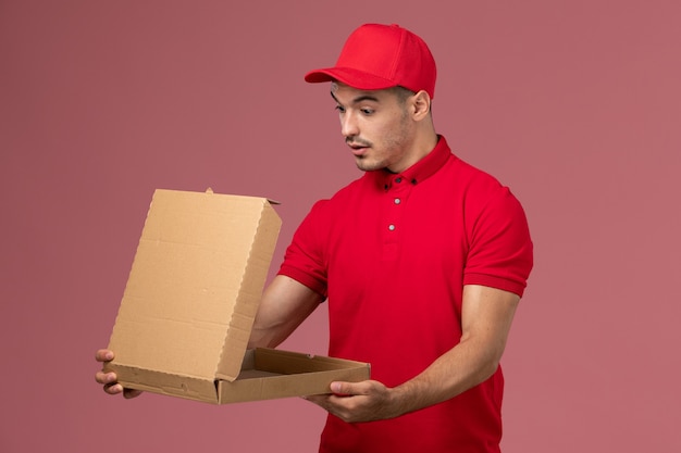 Vooraanzicht mannelijke koerier in rood uniform en cape met voedselleveringsdoos op roze muur levering mannelijke uniform werknemer