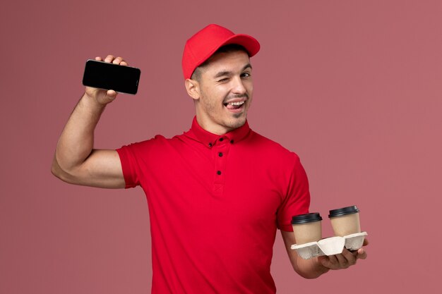 Vooraanzicht mannelijke koerier in rood uniform met bruine bezorgkoffiekopjes en telefoon op het lichtroze muurmannetje