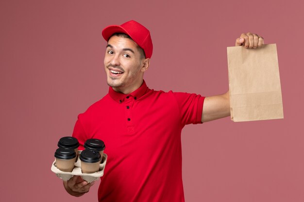 Vooraanzicht mannelijke koerier in rood uniform met bruine levering koffiekopjes met voedselpakket op de roze muur