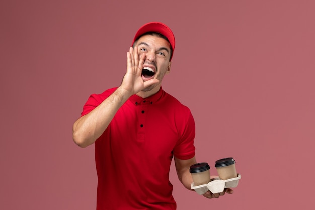 Vooraanzicht mannelijke koerier in rood uniform met bruine levering koffiekopjes schreeuwen op de roze muur mannelijke werknemer