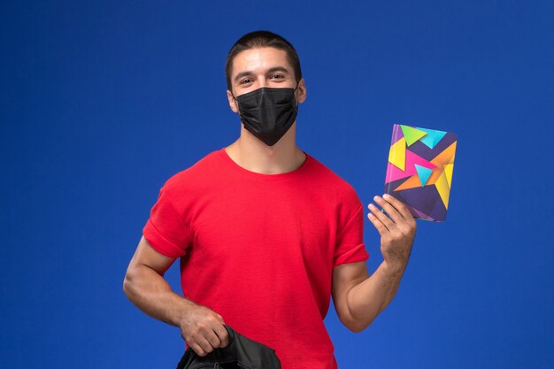 Vooraanzicht mannelijke student in rode t-shirt dragen masker en houdt zijn rugzak en voorbeeldenboek op blauwe achtergrond.