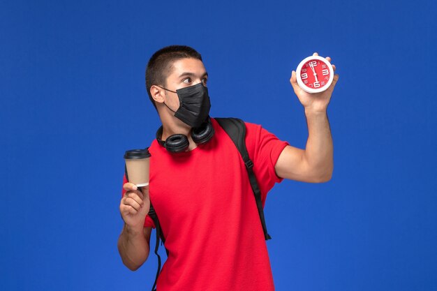 Vooraanzicht mannelijke student in rode t-shirt dragen masker met rugzak houden klokken koffie op blauw bureau.