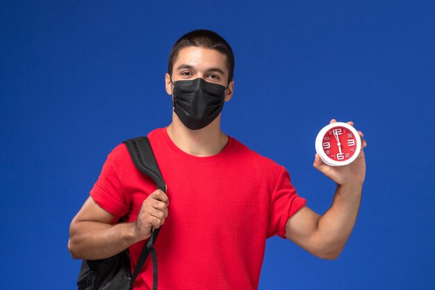 Vooraanzicht mannelijke student in rode t-shirt dragen rugzak met masker houden klokken op de blauwe achtergrond.