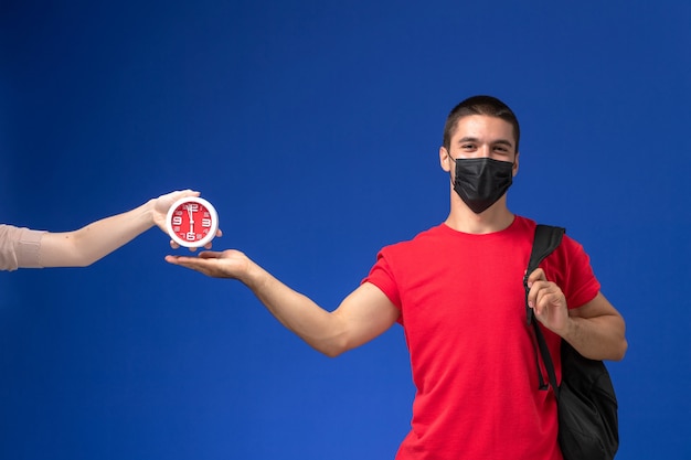 Vooraanzicht mannelijke student in rode t-shirt dragen rugzak met masker poseren op het blauwe bureau.