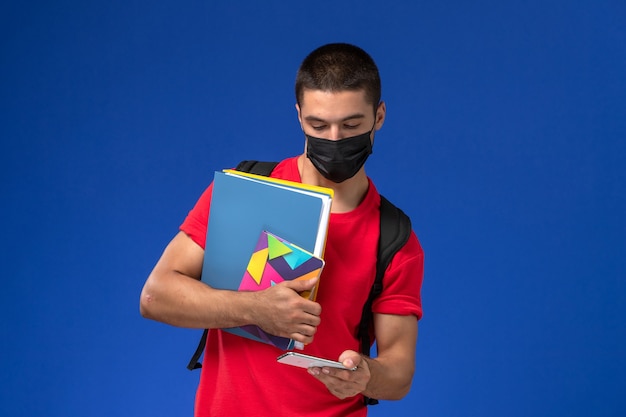 Vooraanzicht mannelijke student in rood t-shirt dragen rugzak met masker bestanden houden en met behulp van zijn telefoon op de blauwe achtergrond.