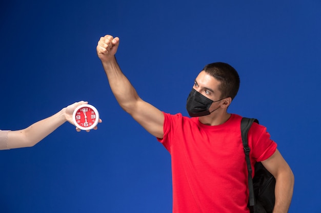 Vooraanzicht mannelijke student in rood t-shirt dragen rugzak met masker poseren op de blauwe achtergrond.