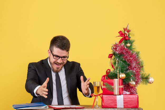 Vooraanzicht mannelijke werknemer achter zijn tafel met cadeautjes en kerstboom op geel