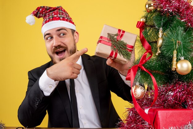 Vooraanzicht mannelijke werknemer achter zijn werkplek met cadeautjes op geel