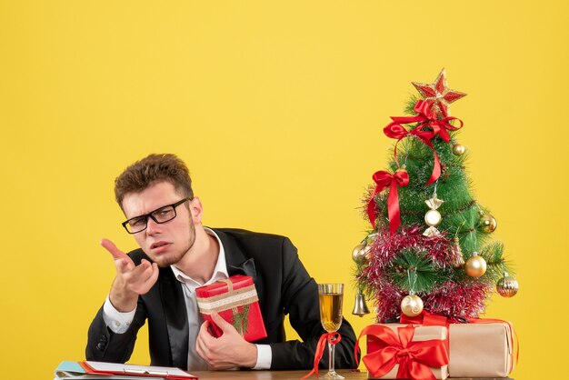 Vooraanzicht mannelijke werknemer achter zijn werkplek met cadeautjes verward op geel