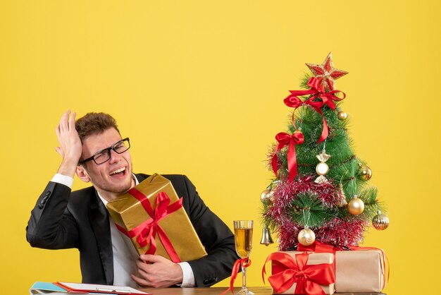 Vooraanzicht mannelijke werknemer achter zijn werkplek met verschillende cadeautjes op geel