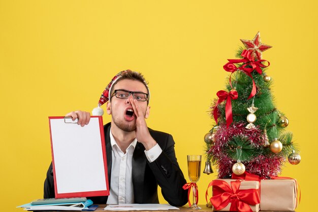 Vooraanzicht mannelijke werknemer met nota rond kleine kerstboom en cadeautjes op geel