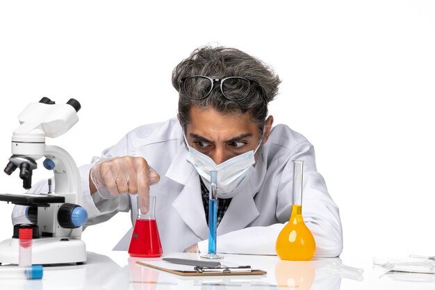 Vooraanzicht middelbare leeftijd wetenschapper in speciaal pak zittend met oplossingen op een wit bureau mannelijke virus wetenschap covid-chemie