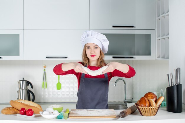 Vooraanzicht mooie jonge vrouw in koksmuts en schort die deeg in de keuken ophouden